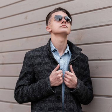 Bir vintage gömlek zarif bir gri ceket moda koyu güneş gözlüğü şık saç modeli ile Yakışıklı genç hipster adam ahşap bir binanın yakınında bir bahar günü sahiptir. Modern adam modeli açık havada rahatlatır.