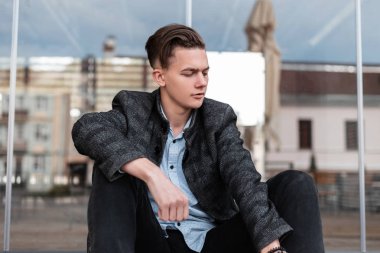 Kot moda gömlek vintage damalı ceket moda saç modeli ile Modern şık genç adam bir bahar gününde cam duvarın yanında oturan dinleniyor. Şehirdeki yakışıklı adam. Bahar stili.