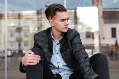 Modaya uygun bir saç modeli nde şık kıyafetler içinde Avrupa moda genç adam şehirde modern bir cam duvar açık yakınında oturan istirahat. İyi adam modeli rahatlatır. Şık erkek giyim.
