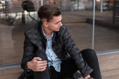 Eski moda gri kareli ceketli, kotlu, şık bir saç stili olan genç, hoş bir adam dışarıda modern cam kapıların yanında oturuyor. Yazlık kafe yakınlarında dinlenen yakışıklı adam. Gençlik tarzı.