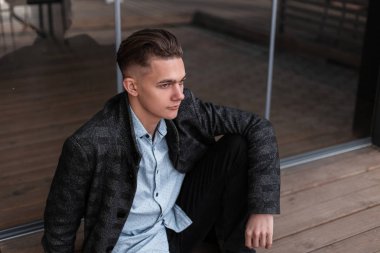 Kot pantolon moda bir gömlek bir vintage ekose ceket şık bir saç modeli ile yakışıklı genç adam bir bahar gününde cam kapı yakınında oturan dinleniyor. Şehirdeki havalı adam hafta sonun tadını çıkarıyor..