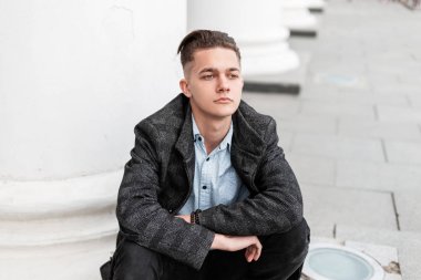 Modaya uygun saç stili olan kotlu şık gömlekli, ekose ceketli genç yakışıklı bir adam klasik bir binanın yanında oturur ve bahar gününün tadını çıkarır. İyi adam şehirde dinleniyor..