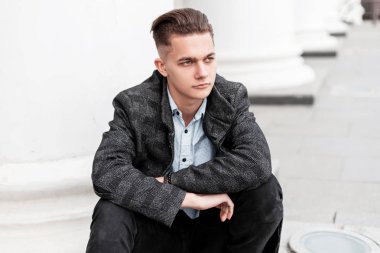 Şık bir saç modeli ile şık bir kot şık gömlek zarif damalı gri ceket modern genç adam şehirde bir vintage binanın yakınında merdivenlerde oturan dinleniyor. Kentsel adam modeli.