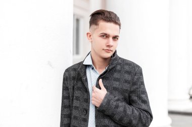 Şık gömlekli, gri, zarif, ekose ceketli, modaya uygun saç stili olan genç yakışıklı bir adam şehirdeki modern bir binanın beyaz sütunlarının yanında duruyor. İş adamı dışarıda modellik yapar..