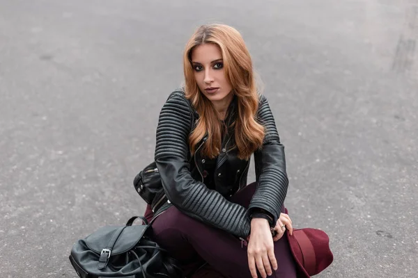 Sexy jeune femme aux cheveux rouges dans une veste en cuir élégant dans un pantalon avec un sac à dos noir à la mode se repose assis sur la route à l'extérieur. Attractive fille européenne aime le week-end. Mode de rue . — Photo
