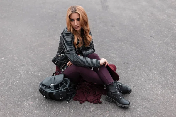 Mulher bonita bonita bonita em uma jaqueta de couro elegante em calças roxas em sapatos pretos vintage com uma mochila elegante senta-se no asfalto na cidade. Menina europeia com cabelo ruivo descansando. Estilo de juventude — Fotografia de Stock