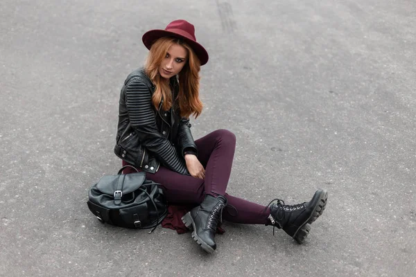 Mulher jovem urbana em um chapéu roxo em uma jaqueta na moda em calças elegantes em botas de couro com uma mochila na moda preta senta-se na estrada na cidade. Menina européia ruiva descansando. Estilo de rua . — Fotografia de Stock