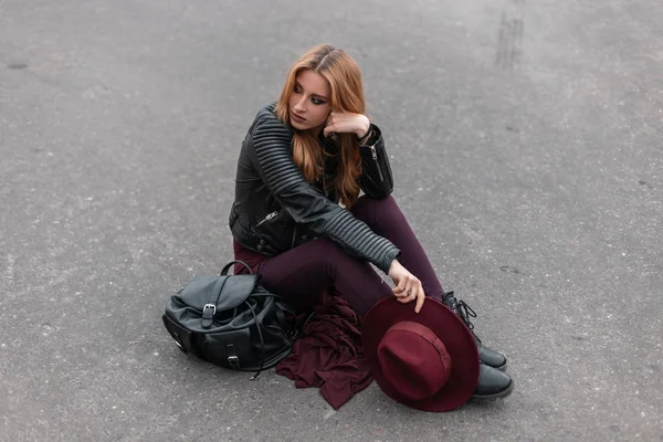 Bella giovane donna con i capelli rossi in una giacca nera in pantaloni viola con cappello vintage in stivali con uno zaino in pelle rilassarsi seduti sull'asfalto sulla strada. Moderna ragazza urbana riposo . — Foto Stock