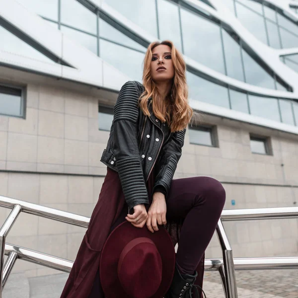 Beautiful pretty young woman in a fashionable leather jacket in purple vintage pants with a hat posing near a modern building in the city. Urban red-haired girl. Street style. — Stock Photo, Image