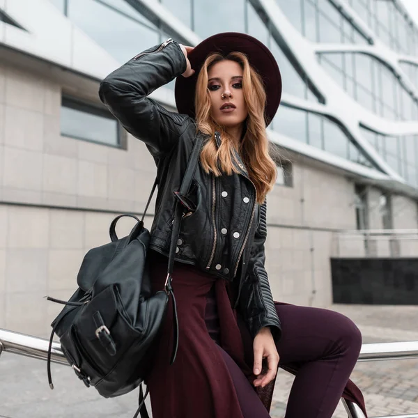 Élégante jeune femme aux cheveux roux dans une veste noire avec un sac à dos en cuir dans un pantalon à la mode avec une cape à la taille est assis sur un tuyau en métal dans la ville près du bâtiment. Fille moderne aime le reste . — Photo