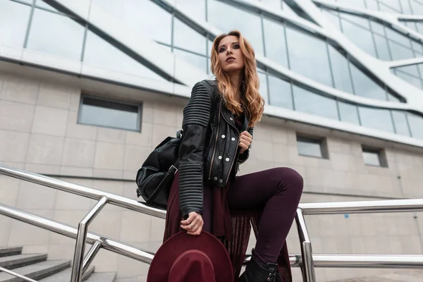 Krásná zrzavá mladá žena s purpurovým kloboukem v módních šatech s koženým batohu leží na kovových trubkách nedaleko moderní budovy ve městě. Evropská dívčí turistka odpočívá. Moderní móda. — Stock fotografie