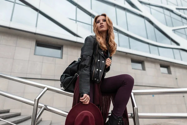 Splendida giovane donna dai capelli rossi con un cappello viola in vestiti alla moda con uno zaino in pelle si siede su tubi metallici vicino a un edificio moderno della città. Ragazza europea turista riposo. Moda moderna . — Foto Stock