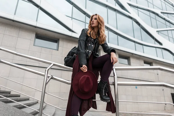 Jolie jeune femme élégante avec un chapeau élégant dans une veste en cuir vintage avec un sac à dos à la mode dans un pantalon élégant dans des bottes assises sur un tuyau métallique près d'un bâtiment moderne. rousse fille repos . — Photo