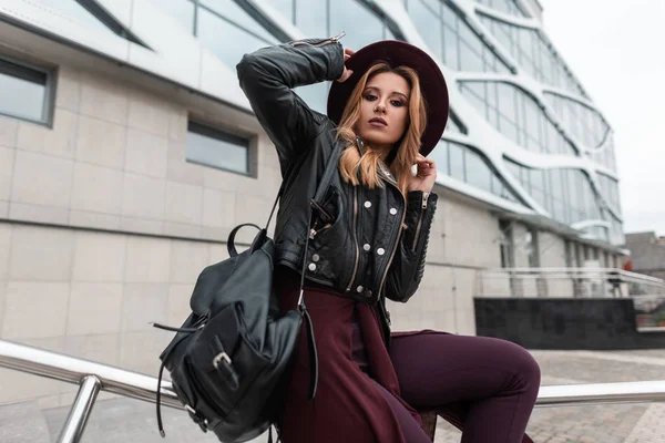 Jovem europeia em um chapéu de luxo em uma jaqueta de couro preto vintage em calças de capa roxa com uma mochila elegante senta-se em um tubo metálico perto de um edifício moderno. Mulher ruiva bonita . — Fotografia de Stock