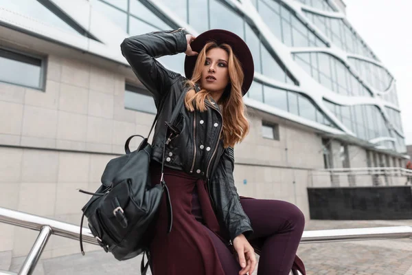 Atractiva joven bonita con un elegante sombrero en una chaqueta de cuero en pantalones vintage con una elegante mochila sentada en tubos de metal cerca de un moderno edificio al aire libre. Chica urbana pelirroja se relaja . —  Fotos de Stock