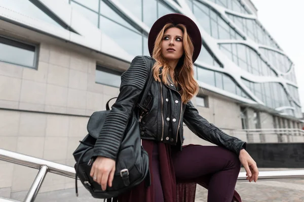 Mulher ruiva muito jovem em um chapéu vintage roxo em uma jaqueta de couro elegante preto com uma mochila na moda em calças da moda perto de um edifício moderno na cidade no dia de primavera. Menina urbana . — Fotografia de Stock