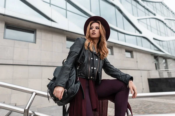 Mulher de cabelos vermelhos jovem moderno em um chapéu chique roxo em uma jaqueta de couro na moda com uma mochila elegante preta em calças da moda perto de um edifício na cidade no dia de primavera. Menina sexy ao ar livre. Moda . — Fotografia de Stock