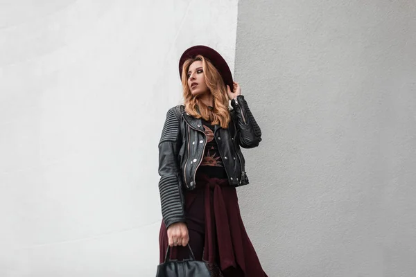 Pretty young woman in a trendy hat in stylish sunglasses in a vintage leather jacket with a backpack with a cape is standing near a white wall in the spring day. Attractive red-haired girl on vacation — Stock Photo, Image