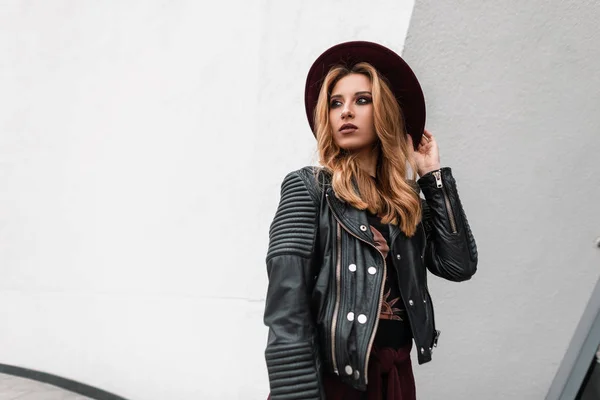 Urbano joven mujer pelirroja bonita en una chaqueta de cuero negro vintage en un elegante sombrero púrpura con hermoso maquillaje está de pie cerca de un edificio moderno en la calle. Chica sexy modelo al aire libre. Primavera . — Foto de Stock