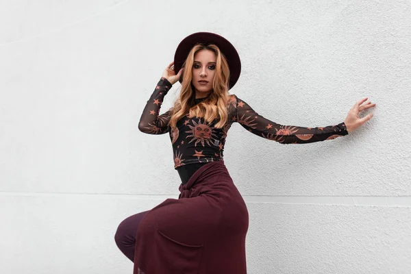 Joven hermosa mujer con estilo en un sombrero de moda en una blusa estampada negro de moda con una capa púrpura posando para un día de primavera cerca de una pared vintage. Chica pelirroja moderna en un paseo. Estilo retro . — Foto de Stock