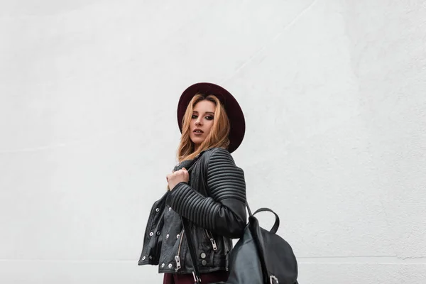 Sexy jeune femme urbaine en chapeau violet élégant dans une veste vintage en cuir noir avec un sac à dos à la mode se tient près d'un mur blanc un jour de printemps. Moderne fille aux cheveux roux modèle à l'extérieur. Style jeunesse . — Photo