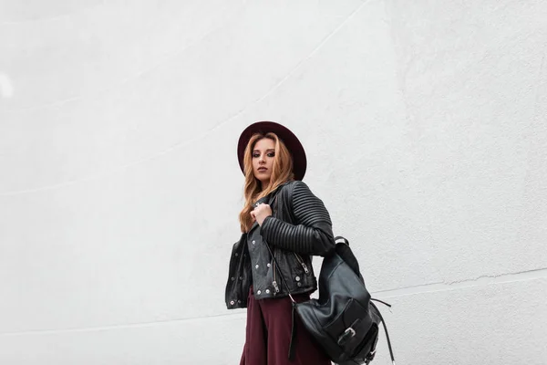 Mulher muito moderna em roupas da moda com uma mochila de couro preto em um chapéu roxo posando perto de uma parede branca vintage ao ar livre no verão. Modelo de menina ruiva muito atraente. Moda . — Fotografia de Stock