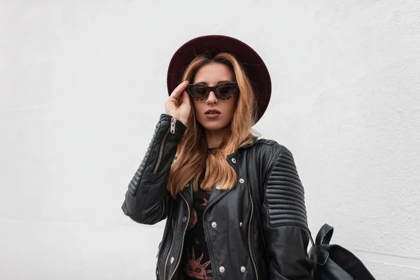 Retrato de una joven hipster sexy pelirroja con un sombrero elegante en gafas de sol oscuras en una chaqueta de cuero de moda con una mochila con un pendiente en la nariz al aire libre cerca de la pared blanca. Chica urbana moderna . —  Fotos de Stock