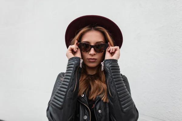 Porträtt av lyxig rödhårig hipster ung kvinna i mörka fashionabla solglasögon i lila hatt i svart snygg skinnjacka nära en vit byggnad utomhus. Vacker modern tjej mode modell. — Stockfoto