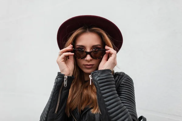 Attractive hipster young woman with sexy lips with beautiful lips in vintage sunglasses in trendy black leather jacket posing near a white vintage wall. Pretty sweet girl. Portrait of a woman's face. — Stock Photo, Image