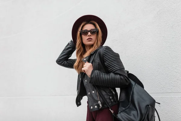 Stylish sexy young hipster woman in purple elegant hat in a vintage leather jacket with a fashionable backpack in trendy sunglasses walks through the city streets. Red-haired pretty girl travels.