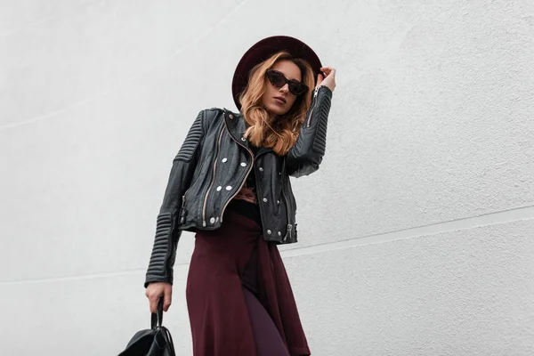 Elégante jeune femme hipster dans un chapeau de luxe en lunettes de soleil dans une veste en cuir dans un pantalon avec un sac à dos avec une cape voyageant dans les rues de la ville près d'un bâtiment blanc. Élégante fille aux cheveux roux . — Photo