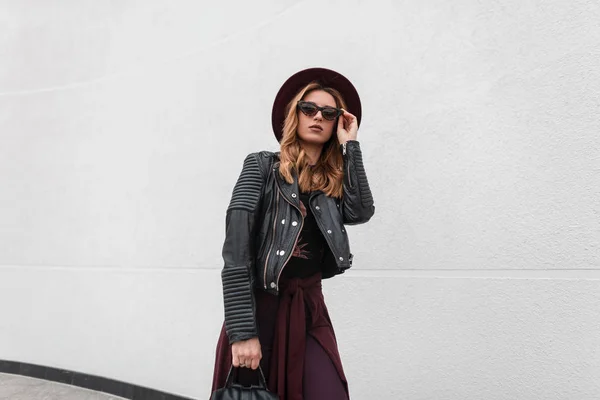 Mujer joven moderna urbana hipster en ropa de moda con una mochila de cuero en un sombrero púrpura posando de pie cerca de una pared vintage al aire libre en verano. Una chica pelirroja muy atractiva. Moda . — Foto de Stock