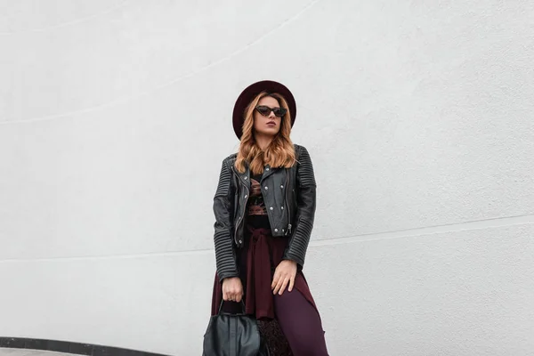 Jolie jeune femme hipster en lunettes de soleil élégantes dans une veste en cuir tendance en pantalon cape avec un sac à dos à la mode dans un chapeau violet près d'un mur blanc vintage dans la rue. Attrayant fille européenne . — Photo