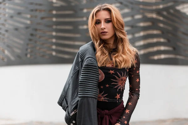 Hermosa mujer joven con el pelo rojo en un pendiente en la nariz en una chaqueta de cuero negro vintage en una blusa de moda con un patrón está de pie en una ciudad cerca de un edificio moderno. Chica bastante elegante . —  Fotos de Stock