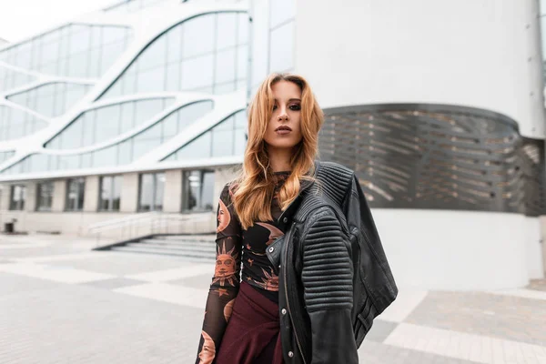 Mujer joven urbana en una blusa estampada de moda con chaqueta de cuero vintage en pantalones con hermoso maquillaje está de pie cerca de un edificio moderno en la calle. Chica sexy con el pelo rojo al aire libre. Primavera . — Foto de Stock