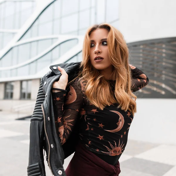 Sexy red-haired young woman with beautiful make-up with an earring in nose in a stylish black leather jacket in a vintage shirt with a pattern of moon and sun poses in a city near a modern building. — Stock Photo, Image