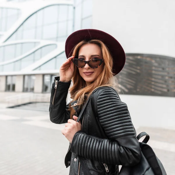 Jolie jolie jeune femme hipster au chapeau violet élégant en lunettes de soleil tendance dans une veste vintage noire avec un sac à dos en cuir se promène dans la ville près d'un bâtiment moderne. Joyeux rousse fille aux cheveux rouges . — Photo