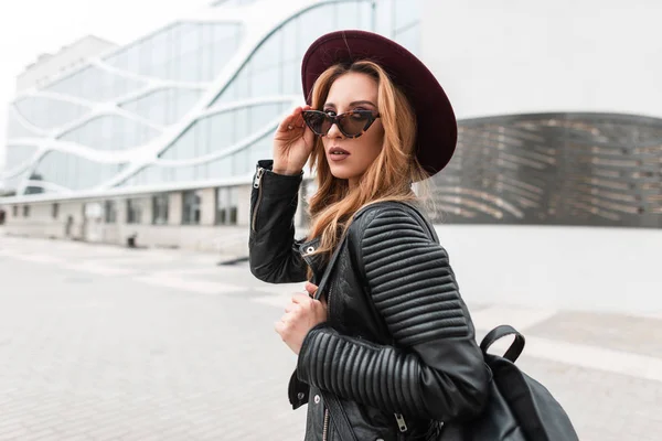 Jolie jeune femme hipster avec un élégant chapeau violet dans des lunettes de soleil élégantes dans une veste en cuir noir avec un sac à dos à la mode se déplace autour de la ville près d'un bâtiment moderne. Européenne fille en plein air . — Photo