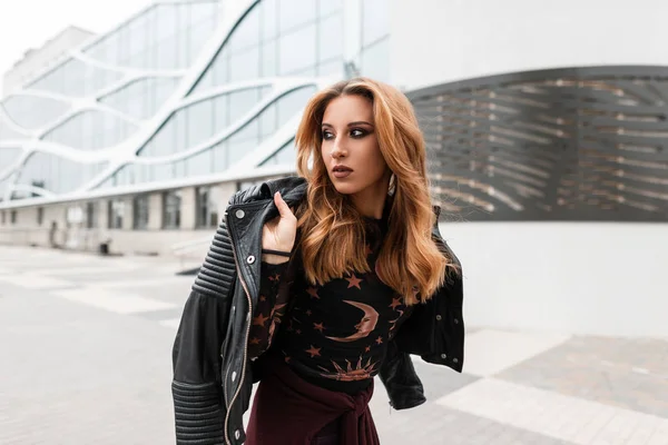 Stylish pretty young woman in a vintage blouse with a pattern in a fashionable black leather jacket with an earring in nose walks down the street near the modern building. Urban red-haired girl. — Stock Photo, Image