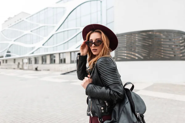 Modern pretty stylish hipster young woman in a fashionable hat in vintage dark sunglasses in a black jacket with a leather backpack is traveling through the city streets. European girl for a walk. — Stock Photo, Image