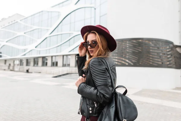 Vacker ung hipster kvinna i mörka solglasögon i en Vintage lila hatt i en skinnjacka med en svart snygg ryggsäck går runt i staden nära en byggnad på vår i dag. Urban Girl resor. — Stockfoto