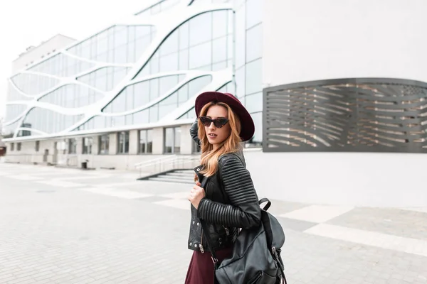 Siyah şık sırt çantası ile bir vintage mor şapka moda güneş gözlüğü Kentsel genç hipster kadın bahar gününde bir binanın yakınında şehir etrafında yürüyor. Pretty kız seyahat. — Stok fotoğraf