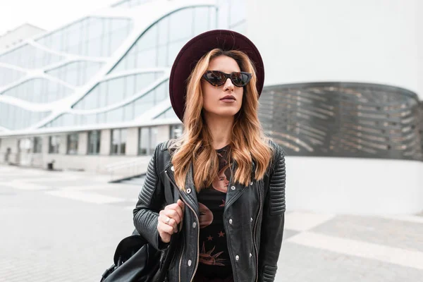 Pretty urban hipster young woman in a fashionable hat in stylish sunglasses in a vintage leather jacket with a backpack walks through the city on a spring day. Attractive red-haired girl travels. — Stock Photo, Image
