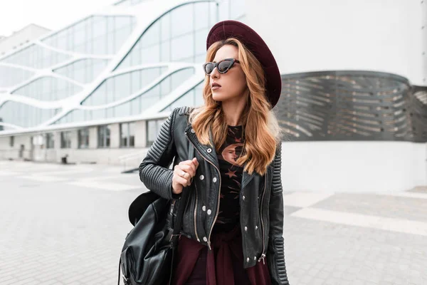 Élégante jolie jeune femme hipster en violet élégant chapeau dans une veste en cuir vintage avec un sac à dos à la mode dans des lunettes de soleil à la mode se promène dans les rues de la ville. Fille touristique aux cheveux roux voyages . — Photo