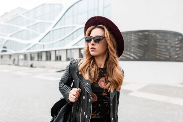 Superbe jeune femme hipster aux cheveux roux avec des lunettes de soleil sombres et un chapeau élégant dans une élégante veste en cuir noir avec un sac à dos tendance dans la ville près d'un bâtiment moderne.Jolie fille européenne pour une promenade — Photo