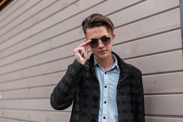 Elegante hombre hipster joven atractivo en gafas de sol oscuras de moda en una chaqueta a cuadros gris con un peinado de moda en una camisa cerca de una pared de madera en la ciudad. Guapo chico modelo disfruta de un paseo . — Foto de Stock
