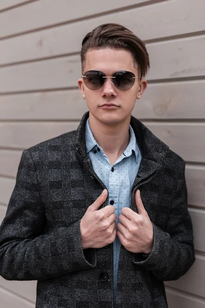 Guapo joven hipster hombre en gafas de sol oscuras en una chaqueta a cuadros gris en una camisa elegante con un peinado de moda posando de pie cerca de una casa de madera vintage. Muchacho europeo moderno.Ropa de moda — Foto de Stock