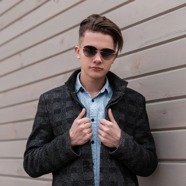Hombre hipster joven guapo serio en gafas de sol de moda en una chaqueta a cuadros en una camisa elegante con un peinado de moda se encuentra cerca de una pared de madera vintage en la ciudad. Buen tipo disfrutando el fin de semana . — Foto de Stock