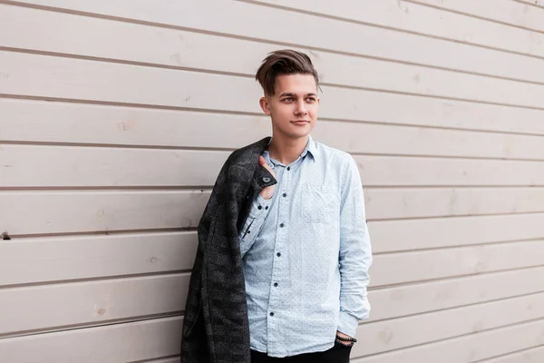 Joven hombre feliz con un peinado elegante con una chaqueta a cuadros gris en una camisa de moda está de pie cerca de una pared de madera en la calle en un día de primavera. Buen chico alegre está disfrutando de un paseo. Prendas masculinas . — Foto de Stock