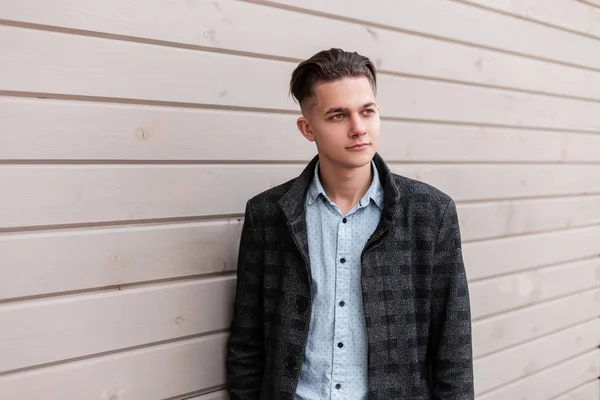 Guapo joven moderno con un peinado en una chaqueta a cuadros gris en una camisa elegante en jeans está de pie cerca de una pared de madera vintage en la ciudad en un día de primavera. Niza europeo chico al aire libre . — Foto de Stock